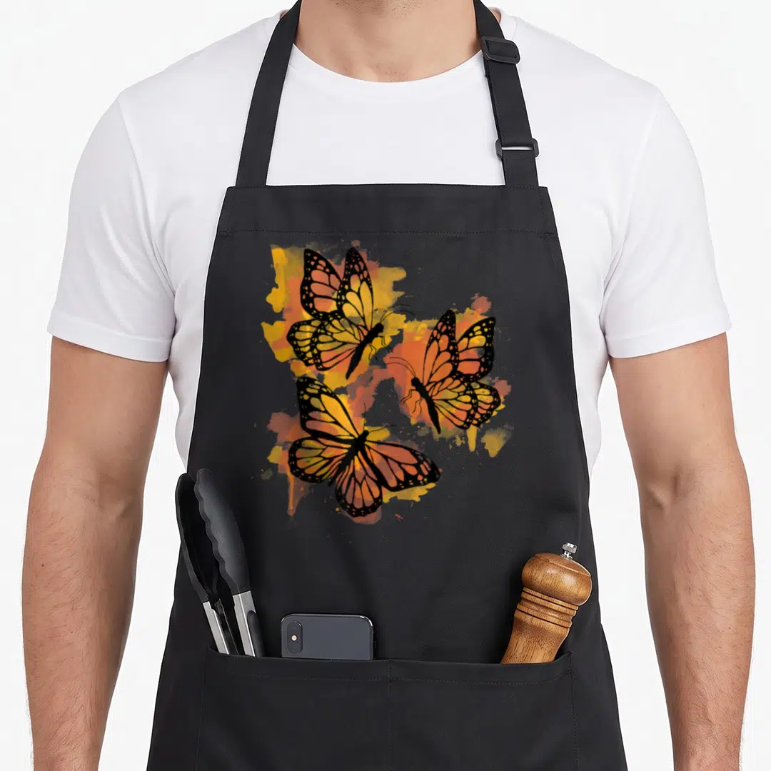 Colourful butterfly apron - Three Monarch butterflies with orange and yellow wings on black background — shown on a black cotton canvas apron against a white background