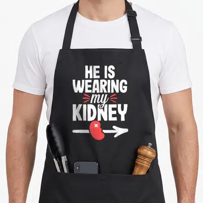 Joke organ apron - White stacked text HE IS WEARING my KIDNEY above a red kidney on a white arrow — shown on a black cotton canvas apron against a white background