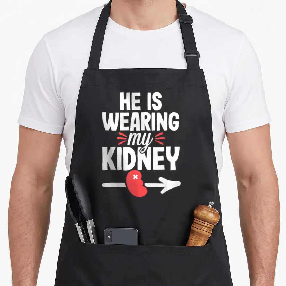 Joke organ apron - White stacked text HE IS WEARING my KIDNEY above a red kidney on a white arrow — shown on a black cotton canvas apron against a white background