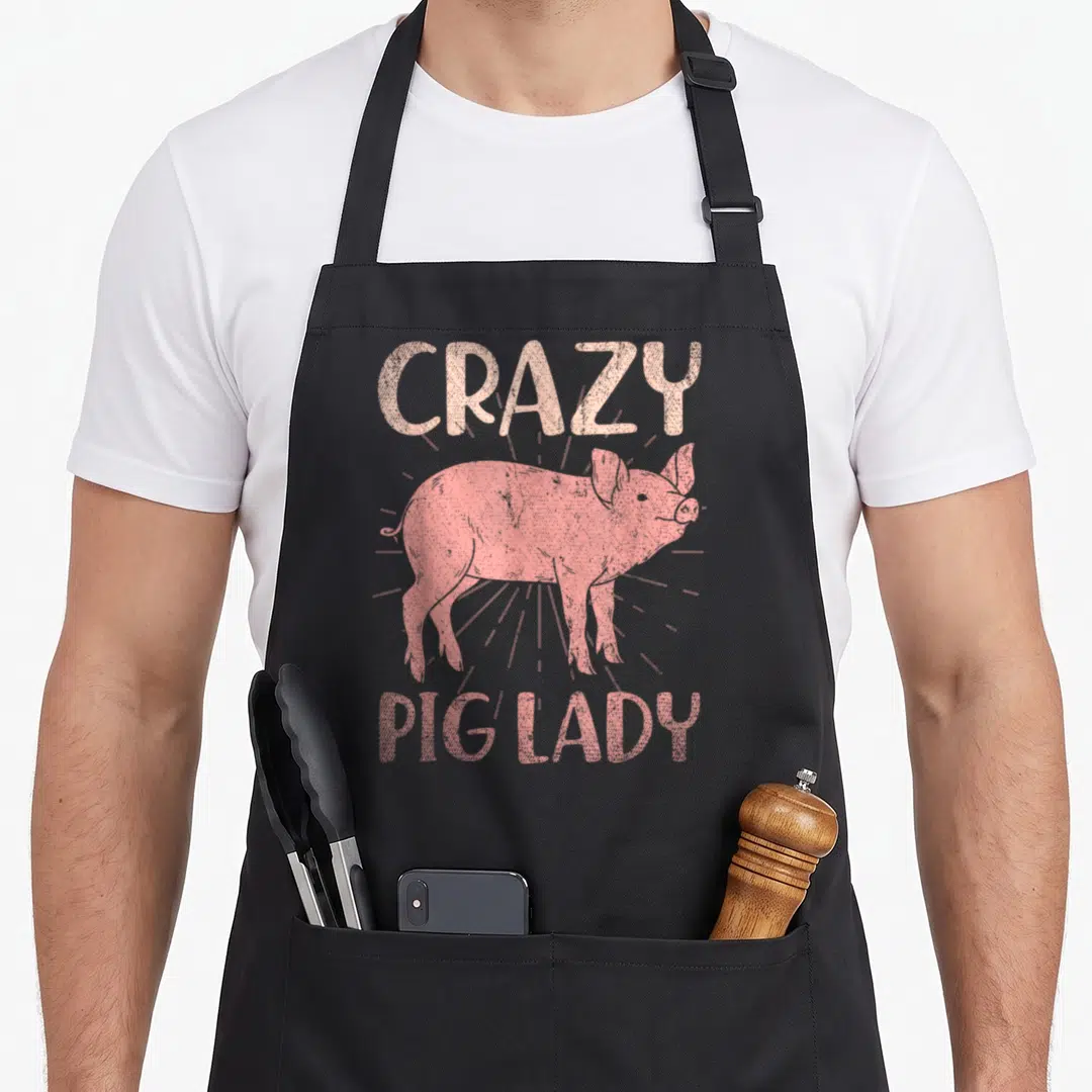 Farmer pig farm apron - Pink pig illustration with CRAZY PIG LADY text and sunburst rays — shown on a black cotton canvas apron against a white background