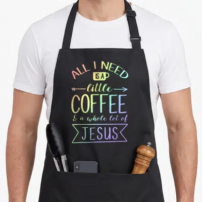 Whole lot coffee apron - Rainbow stacked text reading All I Need Is A Little Coffee And A Whole Lot Of Jesus with cup — shown on a black cotton canvas apron against a white background