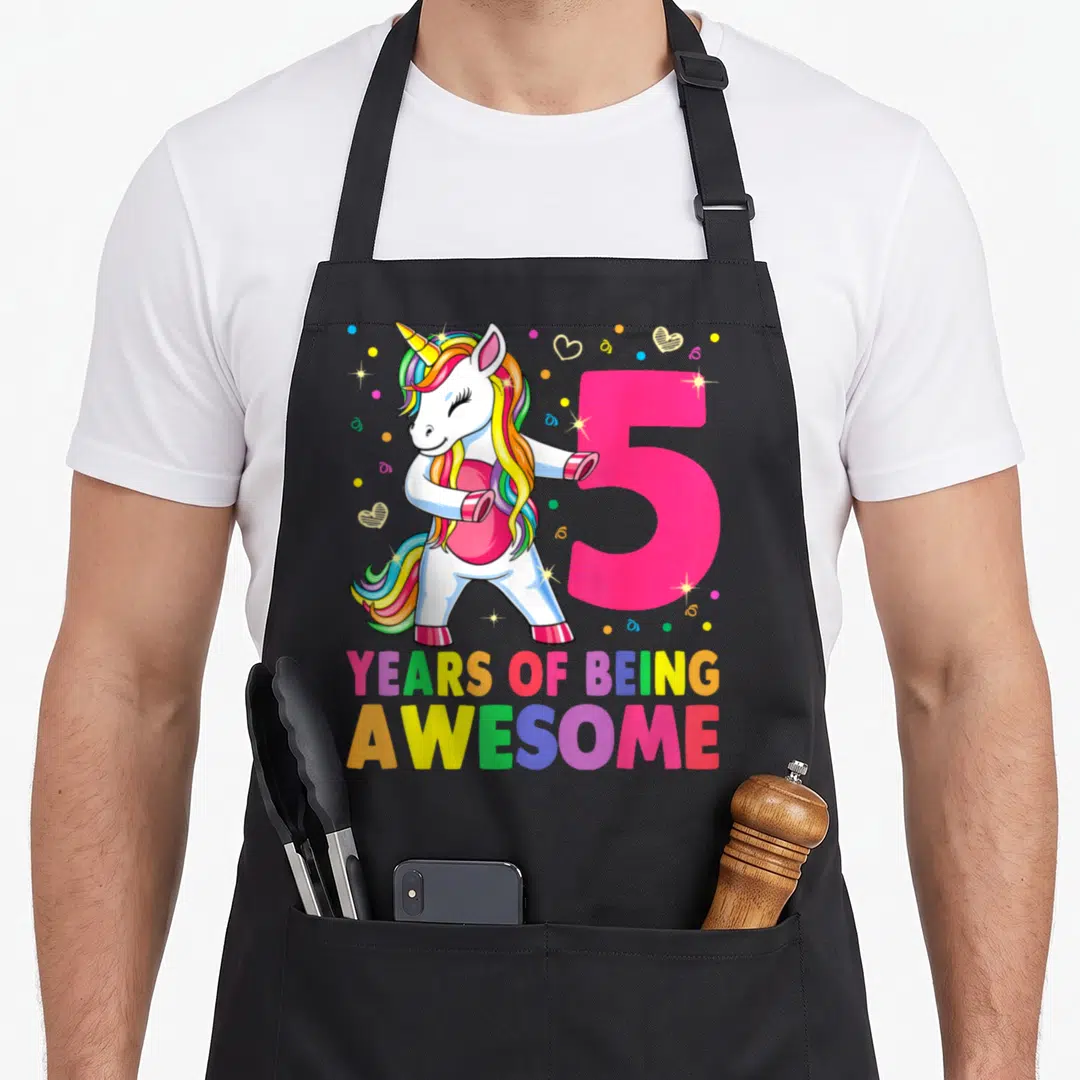 Flossing birthday apron - Cartoon unicorn flossing next to pink number 5 and rainbow text YEARS OF BEING AWESOME — shown on a black cotton canvas apron against a white background