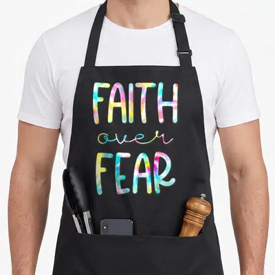 Christian tie-dye apron - FAITH over FEAR text in pink, yellow, blue, and green tie-dye font — shown on a black cotton canvas apron against a white background