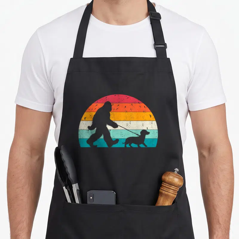 Sasquatch wiener apron - Retro bigfoot silhouette walking a dachshund against a striped sunset — shown on a black cotton canvas apron against a white background