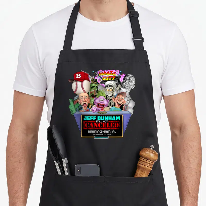 Dunham birmingham apron - Jeff Dunham puppets on blue stage with Magic City text and baseball — shown on a black cotton canvas apron against a white background
