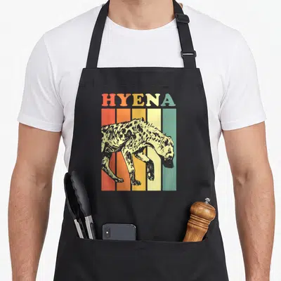 Retro hyena apron - HYENA text above a spotted hyena walking across five vertical stripes in red, orange, yellow — shown on a black cotton canvas apron against a white background