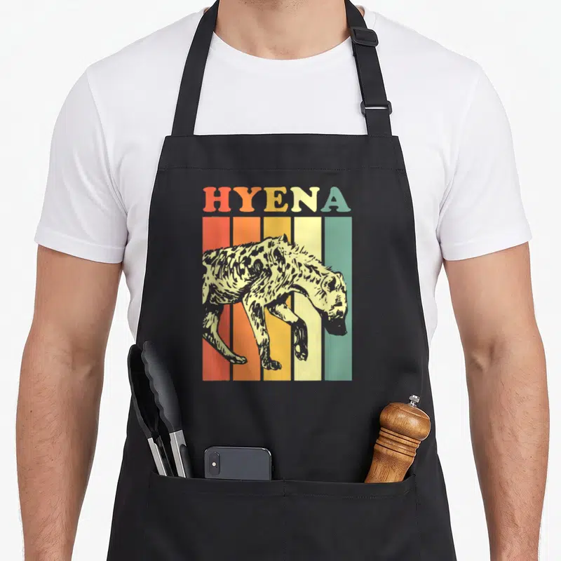 Retro hyena apron - HYENA text above a spotted hyena walking across five vertical stripes in red, orange, yellow — shown on a black cotton canvas apron against a white background