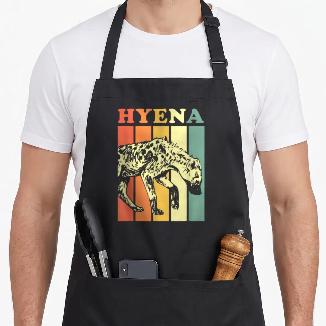 Retro hyena apron - HYENA text above a spotted hyena walking across five vertical stripes in red, orange, yellow — shown on a black cotton canvas apron against a white background