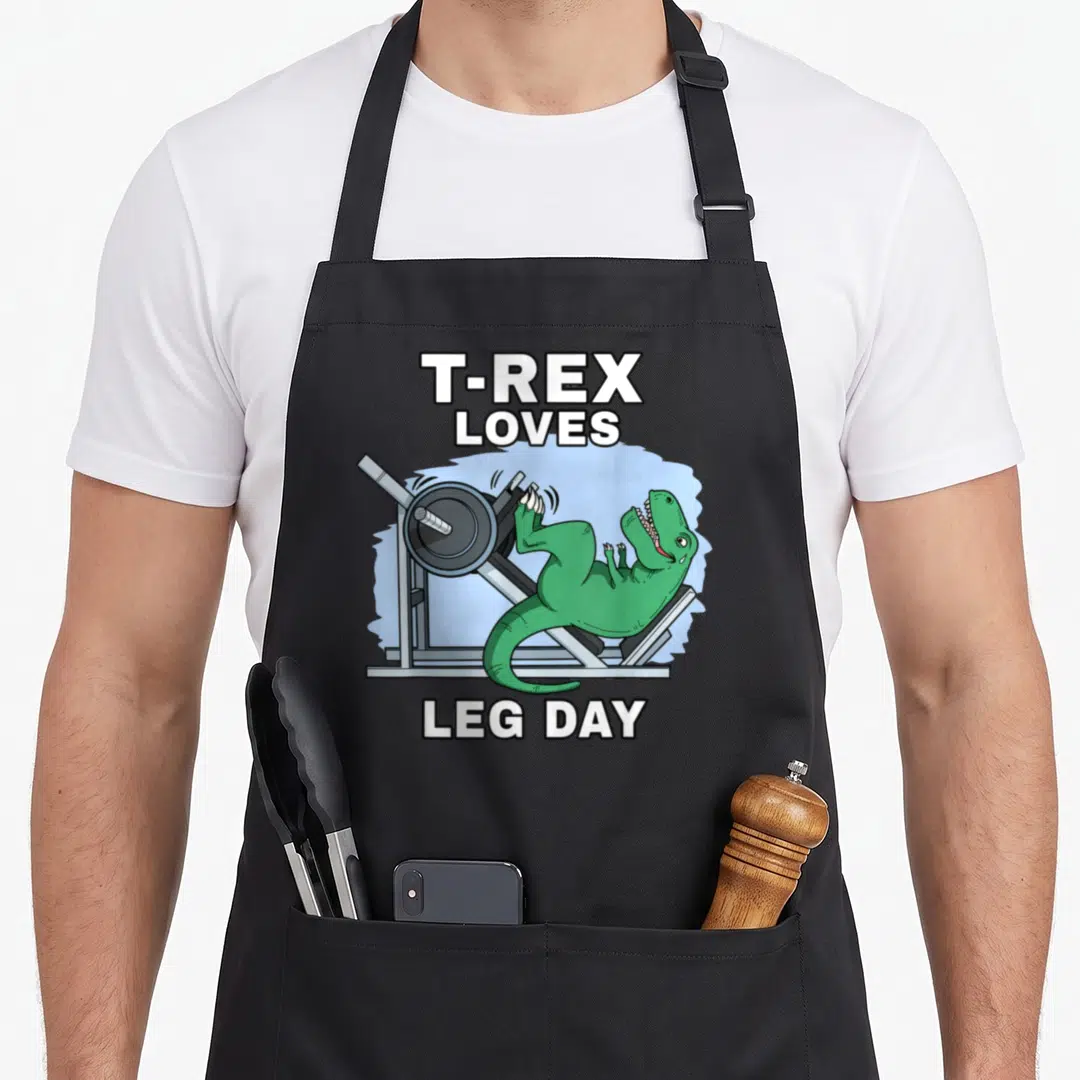 Trex leg apron - Green T-Rex on leg press machine with T-REX LOVES LEG DAY text — shown on a black cotton canvas apron against a white background