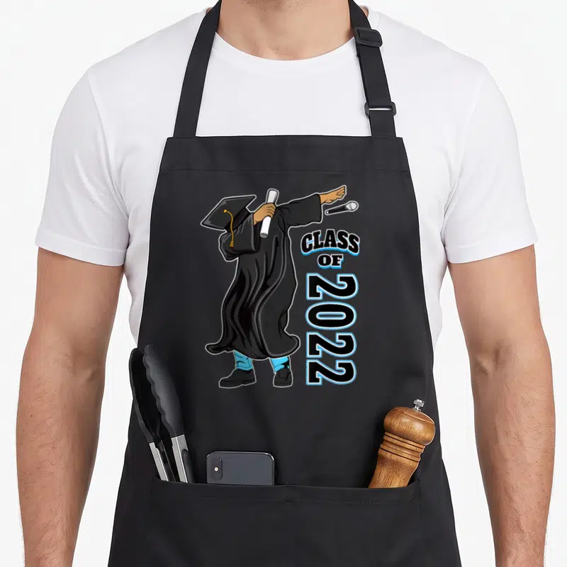 Matter grad apron - Graduate in black gown dabbing holding diploma with vertical CLASS OF 2022 text and blue outlines — shown on a black cotton canvas apron against a white background