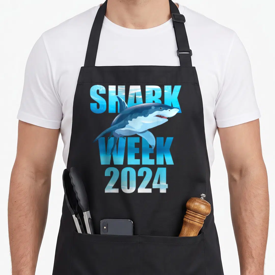 Week family aprons - Cartoon shark swimming over blue SHARK WEEK 2024 water-textured text on black background — shown on a black cotton canvas apron against a white background