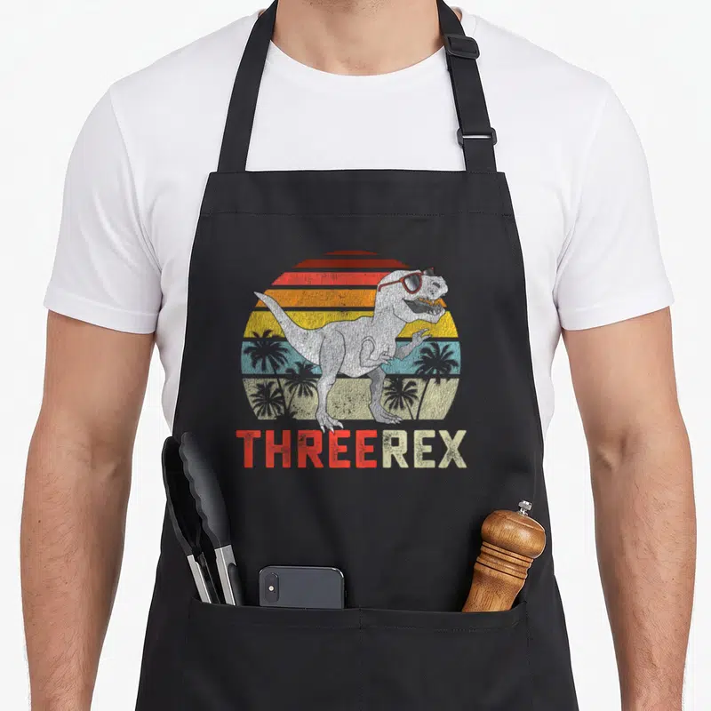Trex t rex apron - T-Rex in sunglasses standing before a striped retro sun with THREEREX text below — shown on a black cotton canvas apron against a white background