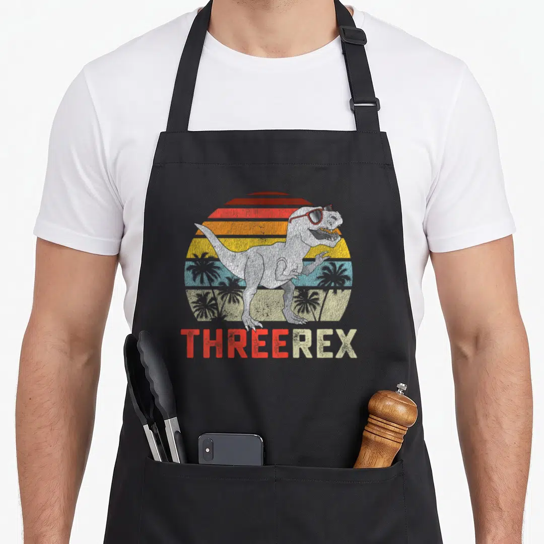 Trex t rex apron - T-Rex in sunglasses standing before a striped retro sun with THREEREX text below — shown on a black cotton canvas apron against a white background