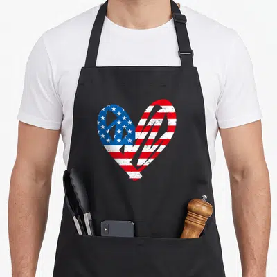 Partiotic 4th of july apron - Distressed heart shape filled with red white and blue USA flag pattern — shown on a black cotton canvas apron against a white background