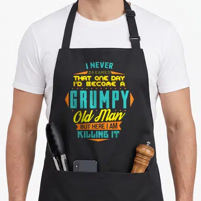 Become never dreamed apron - Stacked teal and yellow text with orange banners reading I never dreamed I'd become — shown on a black cotton canvas apron against a white background