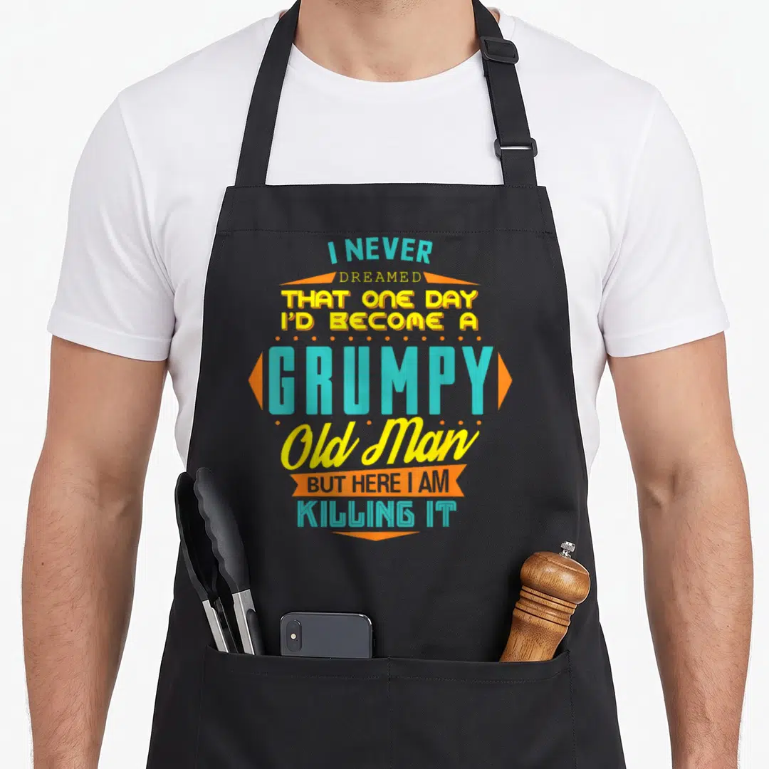 Become never dreamed apron - Stacked teal and yellow text with orange banners reading I never dreamed I'd become — shown on a black cotton canvas apron against a white background