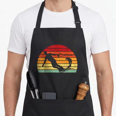 Retro wakeboard wakeboarding apron - Grey water skier silhouette over red orange yellow blue and grey striped sunset sun — shown on a black cotton canvas apron against a white background