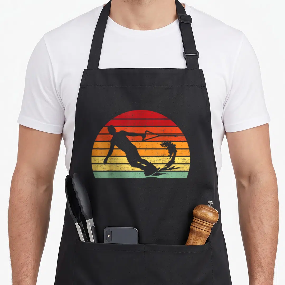 Retro wakeboard wakeboarding apron - Grey water skier silhouette over red orange yellow blue and grey striped sunset sun — shown on a black cotton canvas apron against a white background