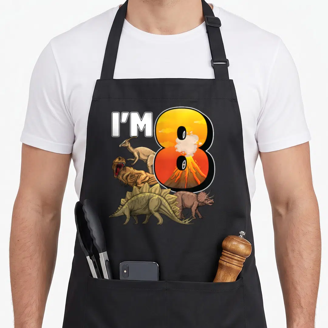 8th dinosaur apron - I'M text beside orange number 8 with volcano and four cartoon dinosaurs in brown green white — shown on a black cotton canvas apron against a white background