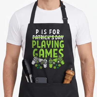 Playing st patrick day aprons - Distressed P Is For Playing Games text with two game controllers and shamrocks in white — shown on a black cotton canvas apron against a white background