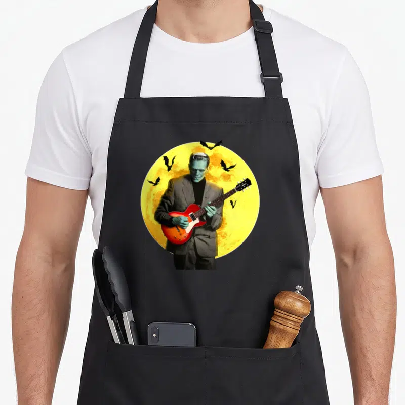 Electric halloween apron - Frankenstein in grey suit holding red guitar in front of yellow moon with black bats — shown on a black cotton canvas apron against a white background