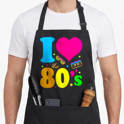 Cute 90s 80s apron - I Love The 80s graphic with pink heart, blue I, cassette tape, shutter shades, and scattered stars — shown on a black cotton canvas apron against a white background