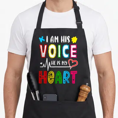 Autism personalized teacher apron - I AM HIS VOICE HE IS MY HEART text with colorful puzzle pieces and heartbeat line — shown on a black cotton canvas apron against a white background