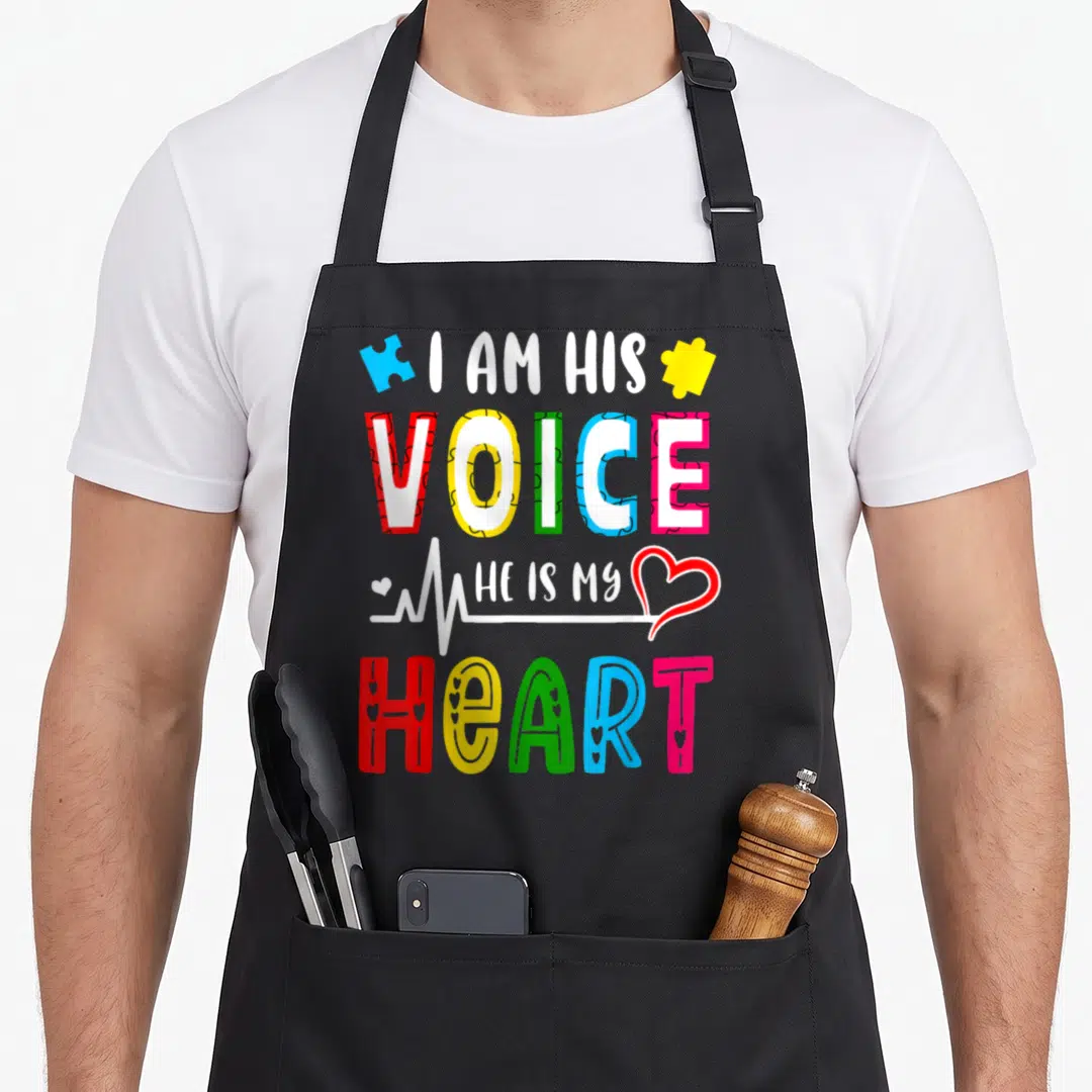 Autism personalized teacher apron - I AM HIS VOICE HE IS MY HEART text with colorful puzzle pieces and heartbeat line — shown on a black cotton canvas apron against a white background