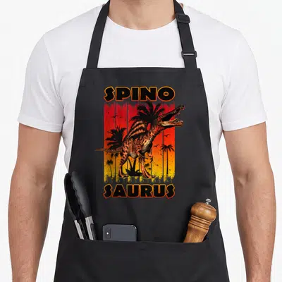 Sunset dinosaur apron - Spinosaurus dinosaur standing before red orange yellow gradient with palm trees and SPINO — shown on a black cotton canvas apron against a white background