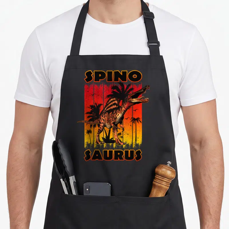 Sunset dinosaur apron - Spinosaurus dinosaur standing before red orange yellow gradient with palm trees and SPINO — shown on a black cotton canvas apron against a white background