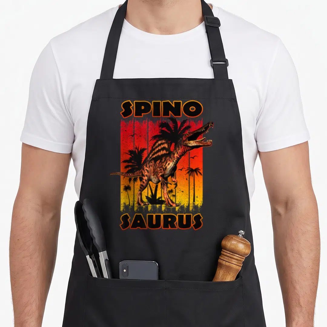 Sunset dinosaur apron - Spinosaurus dinosaur standing before red orange yellow gradient with palm trees and SPINO — shown on a black cotton canvas apron against a white background