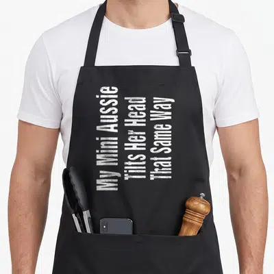 Joke australian aprons - White stacked text reading My Mini Aussie Tilts Her Head That Same Way — shown on a black cotton canvas apron against a white background