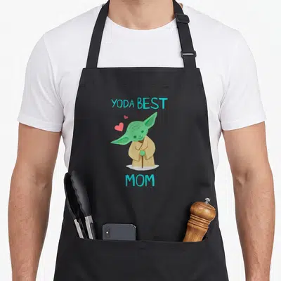 Yoda mother's day aprons - Cartoon green character holding cane with YODA BEST MOM text and two pink hearts — shown on a black cotton canvas apron against a white background