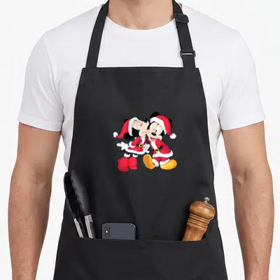 Santa mouse holiday apron - Mickey and Minnie Mouse in red Santa suits kissing in cartoon style with yellow shoes — shown on a black cotton canvas apron against a white background