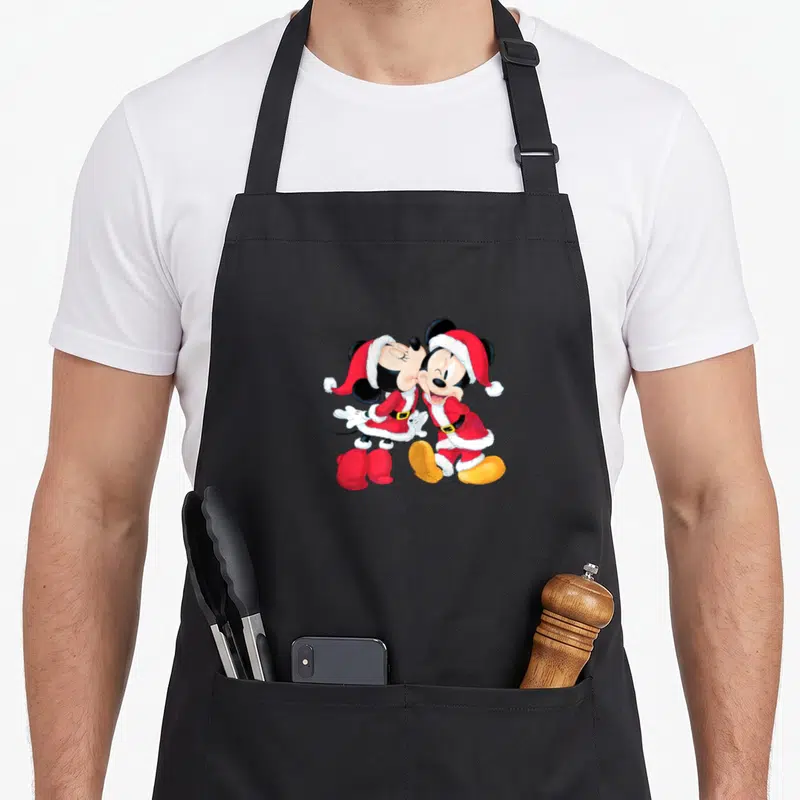 Santa mouse holiday apron - Mickey and Minnie Mouse in red Santa suits kissing in cartoon style with yellow shoes — shown on a black cotton canvas apron against a white background