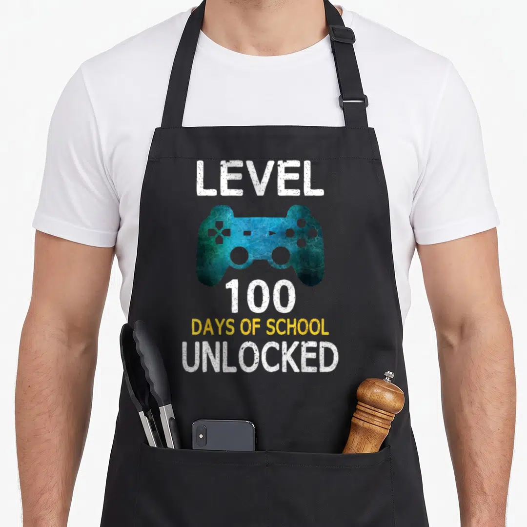 School unlocked video game apron - Blue game controller centered between Level 100 Days of School Unlocked text — shown on a black cotton canvas apron against a white background