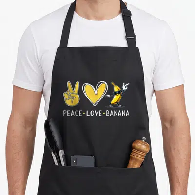 Peace banana apron - Cartoon banana with sunglasses, peace sign, and heart with text PEACE LOVE BANANA — shown on a black cotton canvas apron against a white background