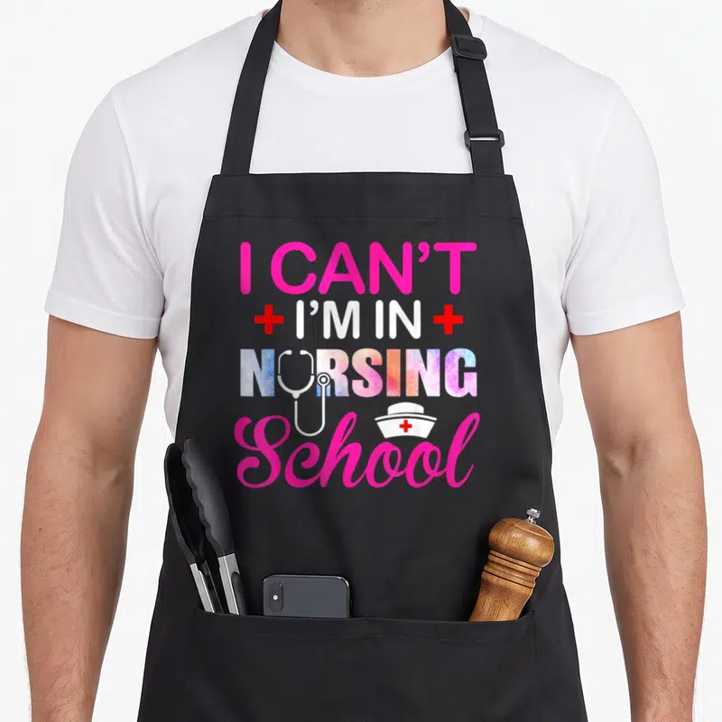 School nurse nursing student apron - I Can't I'm In Nursing School text with stethoscope and nurse hat in pink and white — shown on a black cotton canvas apron against a white background