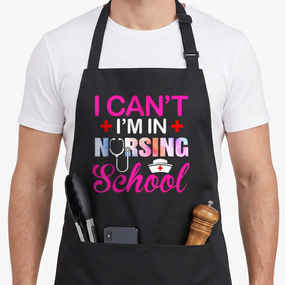School nurse nursing student apron - I Can't I'm In Nursing School text with stethoscope and nurse hat in pink and white — shown on a black cotton canvas apron against a white background