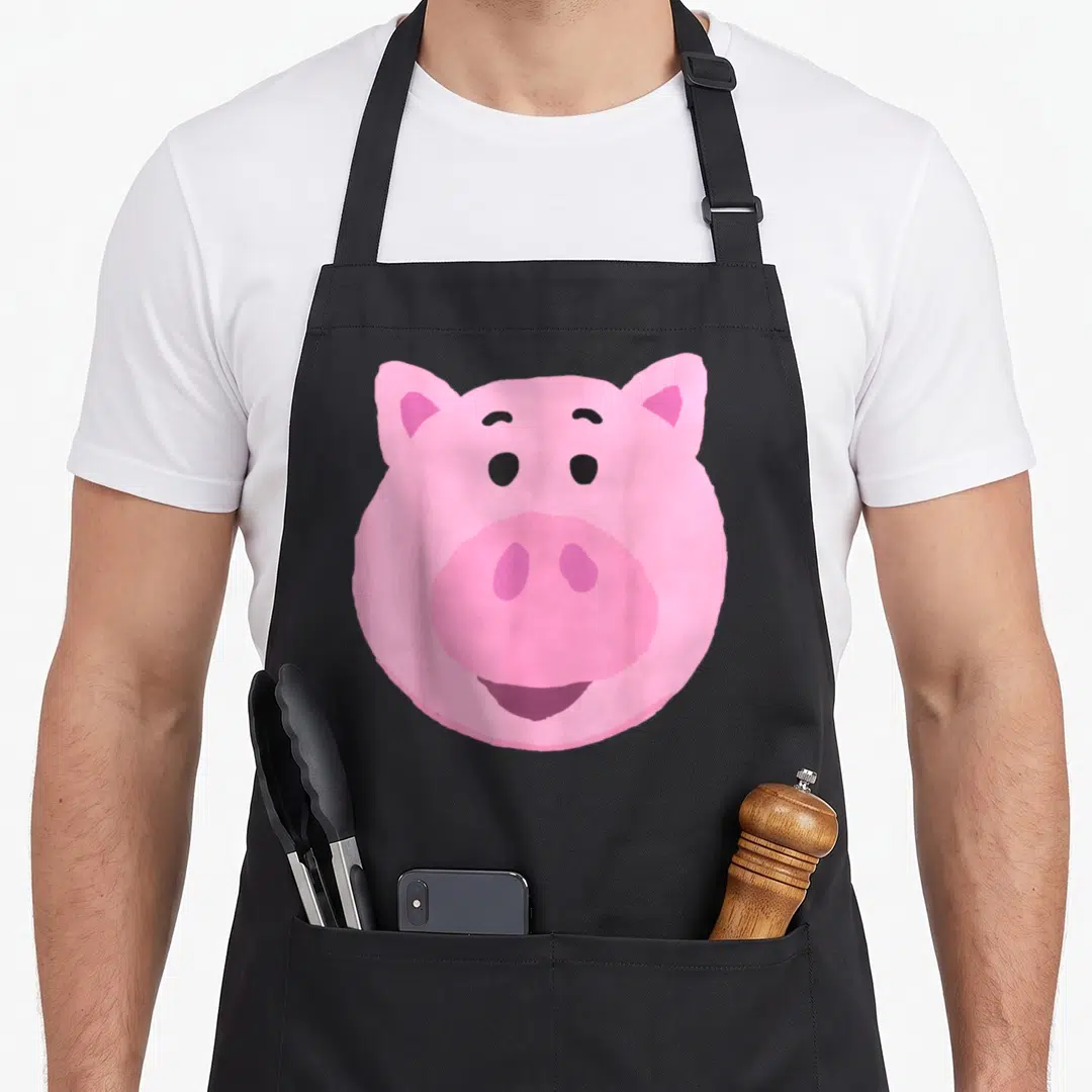 Hamm dog face apron - Pink cartoon pig head with ears eyes and snout centered on fabric — shown on a black cotton canvas apron against a white background