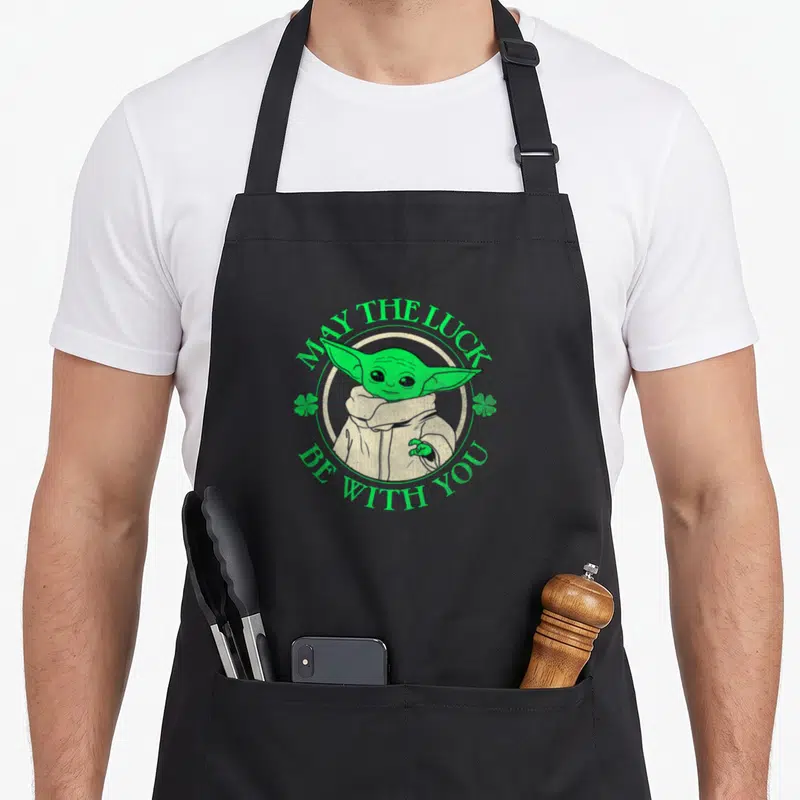 May luck st patrick day aprons - Green Grogu inside white circle with green text MAY THE LUCK BE WITH YOU and shamrocks — shown on a black cotton canvas apron against a white background