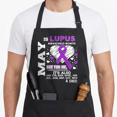 May lupus awareness apron - Purple ribbon over sketched white heart with text listing all twelve months of the year — shown on a black cotton canvas apron against a white background