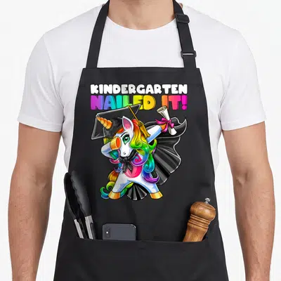 Kindergarten unicorn aprons - Dabbing unicorn in graduation cap holding diploma with rainbow text reading KINDERGARTEN — shown on a black cotton canvas apron against a white background