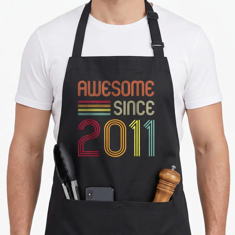 Retro 13th birthday apron - Striped retro typography reading AWESOME SINCE 2011 in orange red yellow and teal — shown on a black cotton canvas apron against a white background