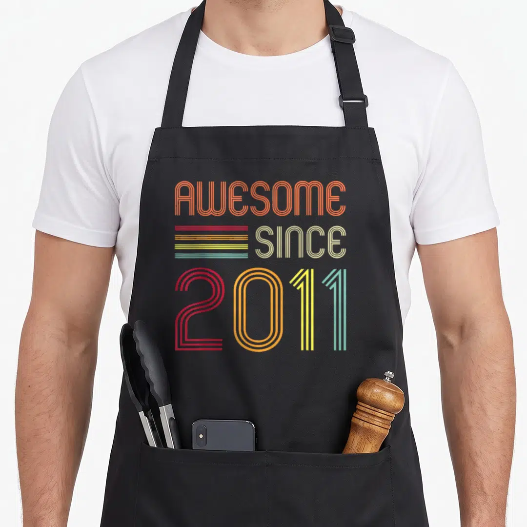 Retro 13th birthday apron - Striped retro typography reading AWESOME SINCE 2011 in orange red yellow and teal — shown on a black cotton canvas apron against a white background
