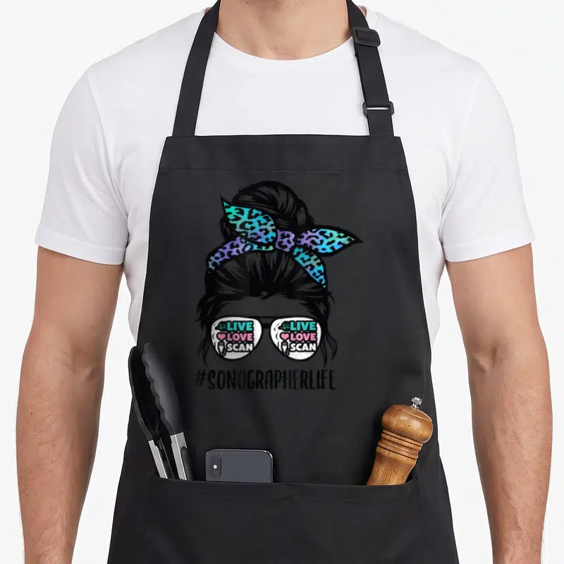 Sonographer messy aprons - Cartoon woman with bun, leopard headband, glasses reading live love scan, and hashtag — shown on a black cotton canvas apron against a white background