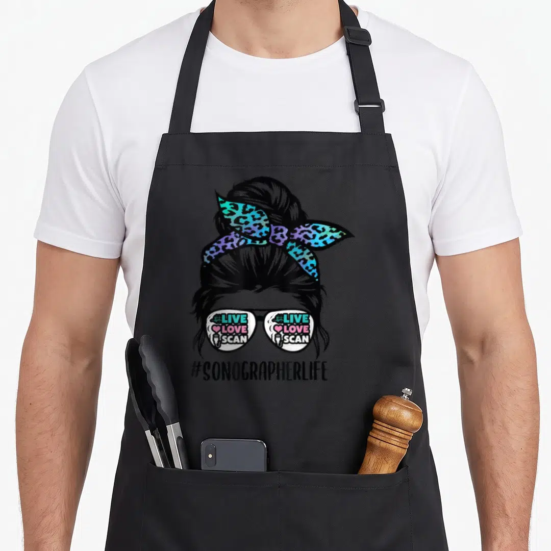 Sonographer messy aprons - Cartoon woman with bun, leopard headband, glasses reading live love scan, and hashtag — shown on a black cotton canvas apron against a white background