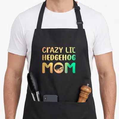 Lil crazy hedgehog lady apron - Stacked text CRAZY LIL HEDGEHOG MOM with hedgehog silhouette in O, orange yellow green — shown on a black cotton canvas apron against a white background