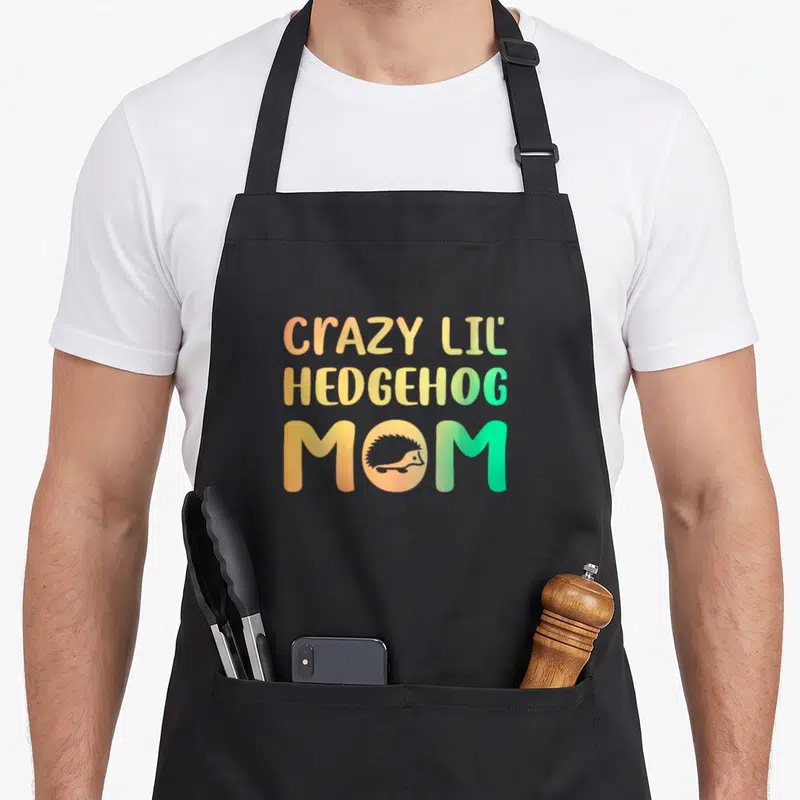 Lil crazy hedgehog lady apron - Stacked text CRAZY LIL HEDGEHOG MOM with hedgehog silhouette in O, orange yellow green — shown on a black cotton canvas apron against a white background
