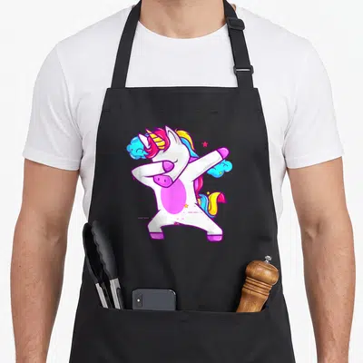 Dab unicorn apron - Cartoon white unicorn dabbing with rainbow mane, blue clouds, and yellow stars — shown on a black cotton canvas apron against a white background